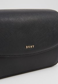 Borsa nera DKNY con esterno testurizzato, dettaglio logo dorato e design elegante. Presenta una chiusura liscia e sottili accenti di cuciture.