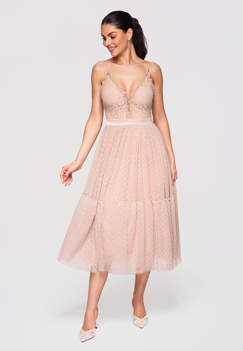 Femme portant une robe midi sans manches, rose poudré, en dentelle et tulle à pois, souriante et regardant vers le bas sur un fond uni.