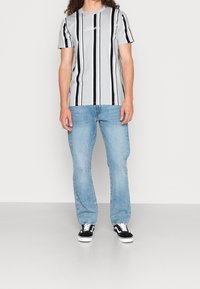 Grå randig t-shirt med svarta vertikala ränder och en vit logotyp på bröstet, kombinerad med ljusblå jeans och svarta sneakers.