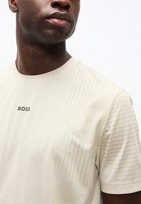 Άνδρας που φοράει μπεζ μπλουζάκι με υφή και το λογότυπο "BOSS" στο κέντρο του στήθους, στραμμένος ελαφρώς προς τα αριστερά μπροστά σε απλό λευκό φόντο.
