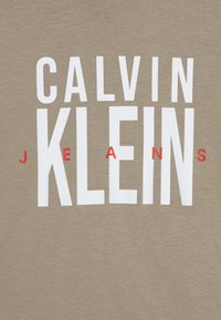 Bež pamučna majica s velikim bijelim slovima koja pišu "CALVIN KLEIN", a "JEANS" je u crvenoj boji, centrirano na prednjoj strani, mekana tekstura.