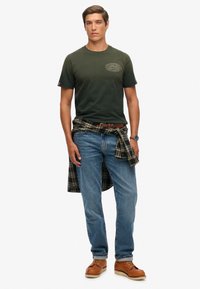 Camiseta de manga corta verde oscuro con un logo, jeans azules, una camisa de cuadros atada alrededor de la cintura y zapatos marrones con cordones.