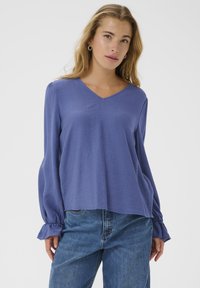 V-neck, long-sleeve blouse in soft, lightweight purple fabric; features gathered cuffs and a relaxed fit. Paired with blue denim jeans.