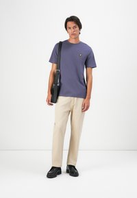 Joven de pie vistiendo una camiseta morada, pantalones beige, zapatos negros y llevando un bolso negro de hombro frente a un fondo blanco liso.
