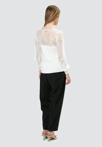 Spitzen- und Strickoberteil mit einem transparenten Spitzenrücken und langen Ärmeln. Vorderseite in reinem Weiß, kombiniert mit schwarzen, lässig geschnittenen Hosen und Riemchen-High Heels.