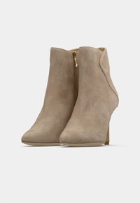 Botas de tobillo de ante beige con punta afilada y detalle de cremallera dorada, que presentan un tacón delgado y una textura suave.