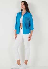 Camicia blu con bottoni, in tessuto textured, con una tasca decorativa ricamata e pantaloni bianchi aderenti abbinati a sneakers.