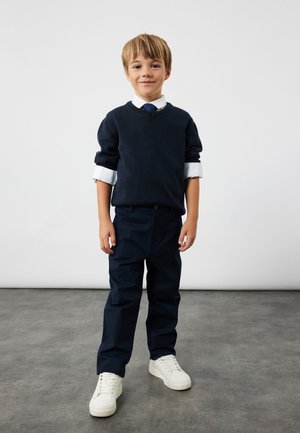 Maglione in misto lana di colore blu navy sopra una camicia bianca con bottoni, accompagnato da una cravatta blu navy. I pantaloni blu navy e le sneakers bianche completano l'outfit.