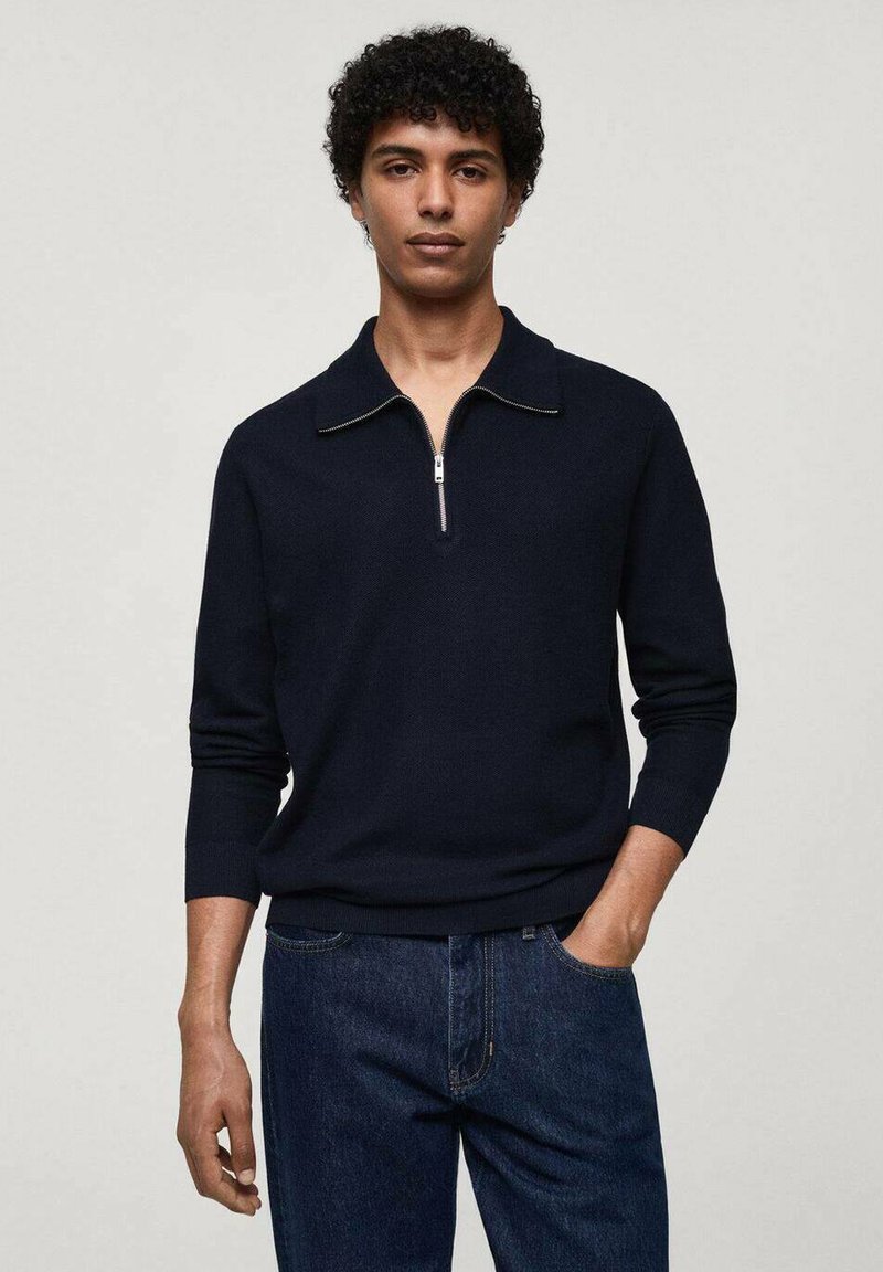 Pulover tricotat bleumarin cu guler cu fermoar, mâneci lungi și mansete cu șnur. Asortat cu blugi denim închis. Textură netedă.