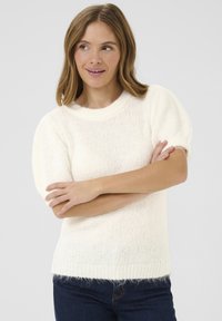 Un pull en tricot de couleur crème avec des manches courtes bouffantes et un col rond. La texture semble douce et fluffy, avec un ourlet côtelé. Un jean en denim est porté.