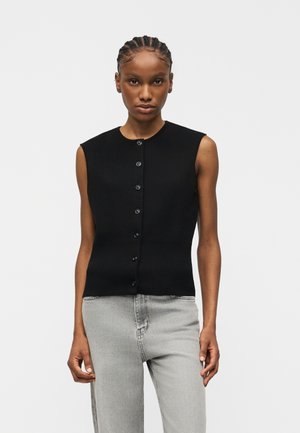 Fonott hajú nő, fekete, ujjatlan, gombos felsőt és világosszürke farmert visel, sima, világos háttér előtt áll.