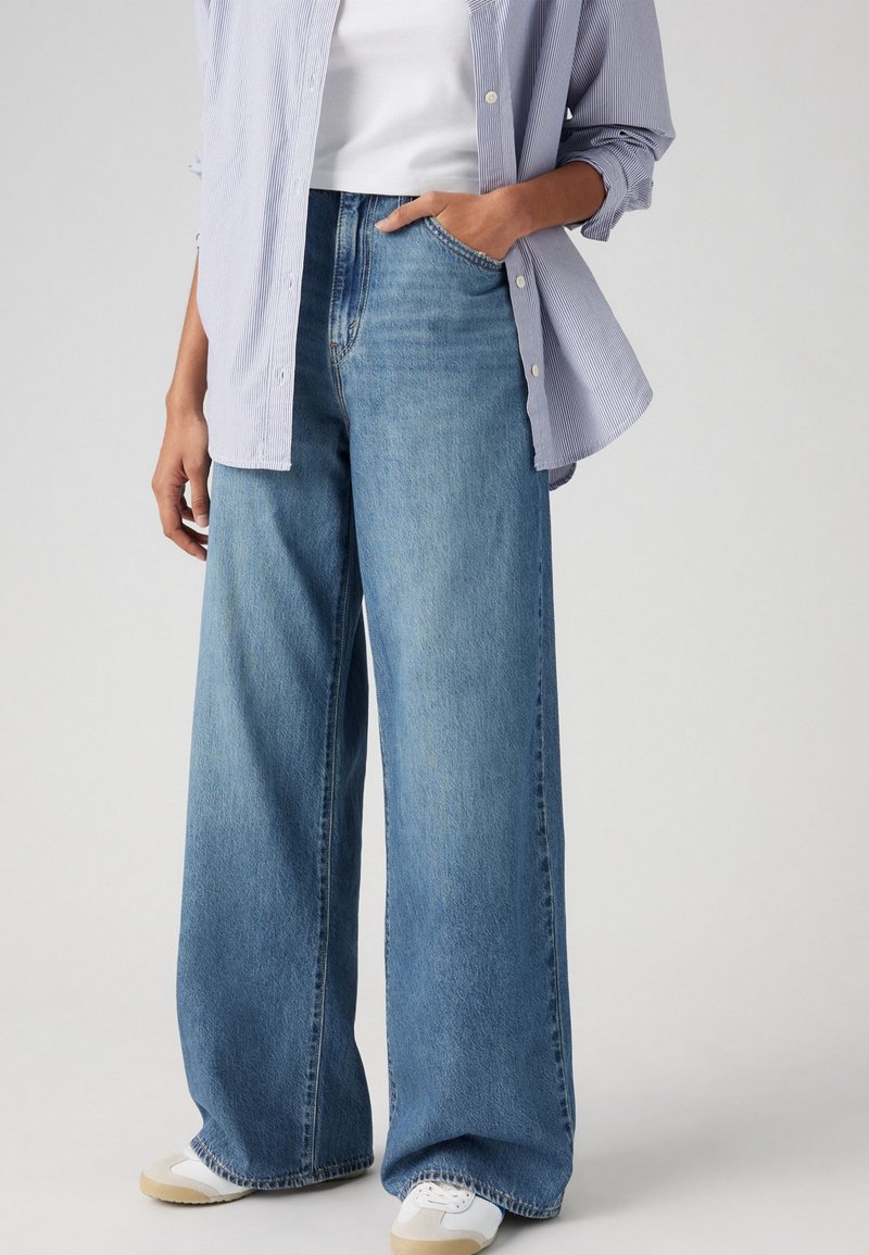 High-waisted, wide-leg blauwe denim jeans met een vervaagd uiterlijk, voorzakken en zichtbare stiksels. G dragen met een lichtblauw gestreept shirt.