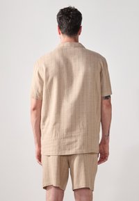 Beige kortermet skjorte og shorts-sett, med et teksturert stoff med vertikale striper og en avslappet passform. Vist fra baksiden.