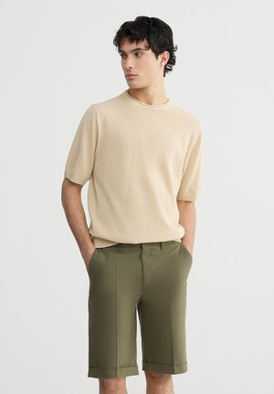 Camicetta corta a maniche corte beige in maglia abbinata a pantaloni corti olive verdi su misura. L'outfit presenta una vestibilità rilassata e una texture morbida.