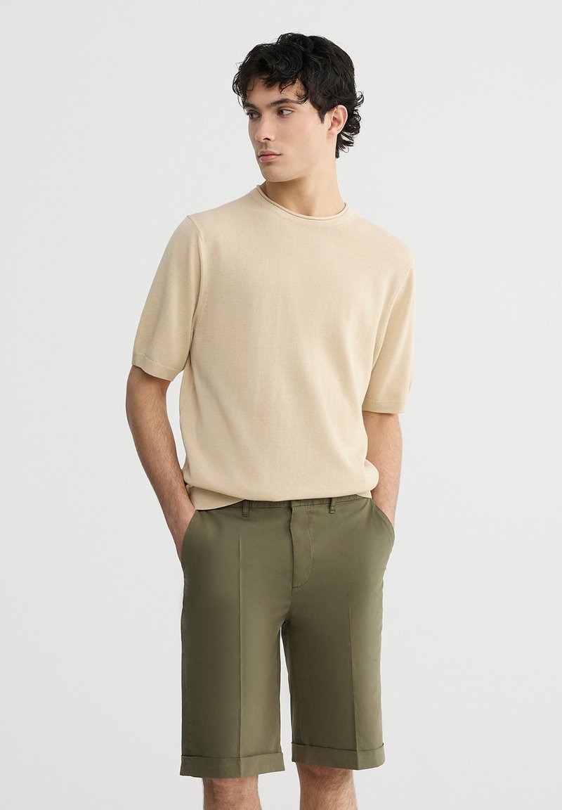 Camicetta corta a maniche corte beige in maglia abbinata a pantaloni corti olive verdi su misura. L'outfit presenta una vestibilità rilassata e una texture morbida.