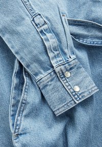 Chemise en denim bleu clair avec un poignet à boutons, présentant un design classique, des coutures visibles et une texture douce.