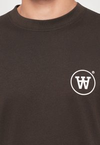 Nahaufnahme einer Person, die ein dunkelbraunes Rundhals-Shirt trägt, das auf der Brust ein weißes, kreisförmiges Logo mit umgekehrten doppelten "A"-Buchstaben zeigt.