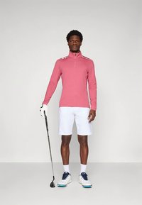 Camisola rosa claro de manga comprida com fecho, combinada com calções brancos. Sapatos de golfe brancos com detalhes azuis. Um taco de golfe segurado na mão esquerda.