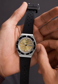 Timex watch with a gold dial, black rotatable bezel, silver stainless steel case, and textured black rubber strap, showing the day and date.