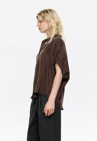 Brown, short-sleeve button-up shirt in a soft, smooth fabric. Loose fit with a rounded hem and subtle side slits. Paired with gray trousers.