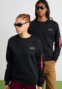 Zwarte sweatshirt met een ronde hals, textuurstof, logo op de voorkant en een gezoomde mouwzak met een rode label. Draagt met een zonnebril.