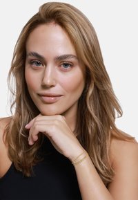 Primo piano di una donna con capelli biondi ondulati di lunghezza spalla, che indossa un top nero. Ha un bracciale d'oro con dettagli delicati a catena al polso.