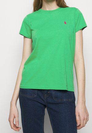 Kobieta ubrana w jasnozieloną koszulkę z krótkim rękawem z małym różowym haftowanym logo na lewej piersi, w połączeniu z ciemnoniebieskimi dżinsami.