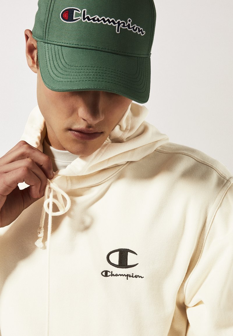 Haut à capuche crème avec cordon de serrage et logo Champion noir brodé. Casquette verte foncée avec le texte Champion brodé sur le devant.