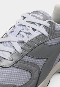 Κοντινό πλάνο λευκού και γκρι αθλητικού παπουτσιού με πλέγμα ύφασμα, λευκά κορδόνια και λογότυπο της εταιρείας στη γλώσσα.