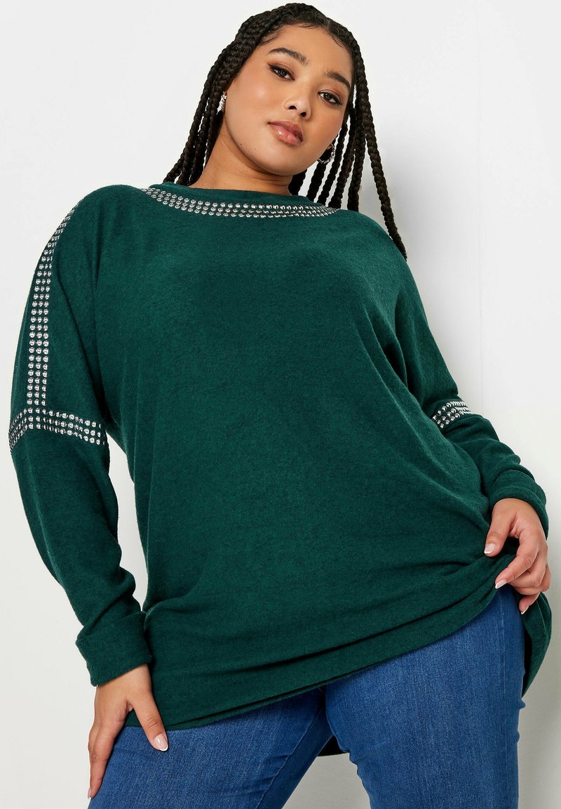 Maglione verde oversize con una consistenza morbida, caratterizzato da una fascia di borchie decorative in argento lungo le spalle. Abbinato a jeans blu.