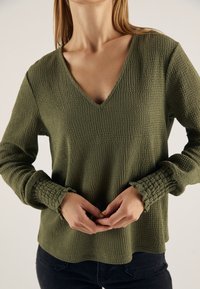 Suéter verde de cuello en V con tejido texturizado, mangas largas con puños fruncidos y un corte relajado. Lleva puesto sobre pantalones oscuros.