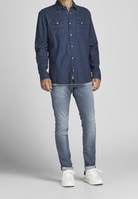 Mörkblå knäppt skjorta med två bröstfickor, kombinerad med ljusblå jeans och vita sneakers, visad mot en neutral bakgrund.