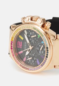 Montre chronographe dorée avec un cadran noir, présentant des marqueurs colorés et une lunette ornée de joyaux dans des teintes de violet, vert et jaune.