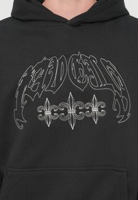 Μαύρο φούτερ με κουκούλα, με λευκή κεντημένη γραφή και λουλουδένια σχέδια. Το υλικό φαίνεται μαλακό, με μια τυπική τσέπη καγκουρό στο μπροστινό μέρος.