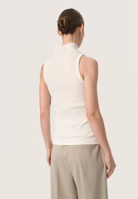 Weißes geripptes ärmelloses Turtleneck mit einer enganliegenden Passform und feiner Textur. Getragen mit beigen Hosen, von hinten betrachtet.