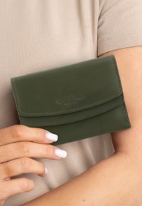 Portafoglio in pelle verde con chiusura a patta, caratterizzato da un logo impresso e bordi cuciti. Tenuto in mano su uno sfondo beige.