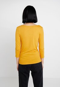 Žlutý těsný dlouhý rukáv top s kulatým výstřihem, vyrobený z měkké směsi bavlny. Nesený s černými kalhotami s vysokým pasem. Pohled zezadu.