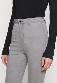 Jeans gris de talle alto con una textura suave, cierre de botón y bolsillos frontales. Combinados con una blusa negra de manga larga con detalles dorados.