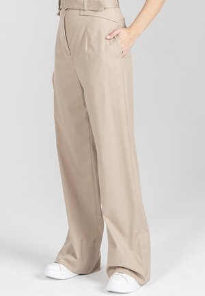 Beżowe szerokie spodnie z wysokim stanem i bocznymi kieszeniami, noszone z białymi trampkami, widoczne na osobie od talii do stóp.