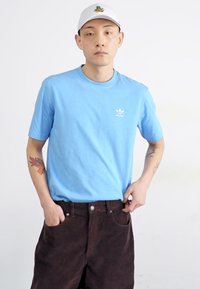 Camiseta de algodón de color azul claro con cuello redondo y pequeño logo blanco, combinada con pantalones de pana marrón oscuro, que cuentan con bolsillos delanteros.