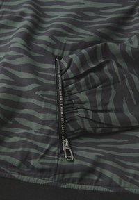 Tissu rayé en noir et vert, avec une poche zippée sur le côté, présentant une surface texturée et des détails élastiques.
