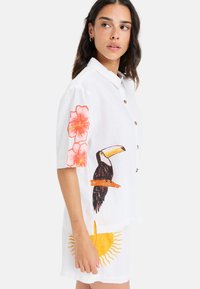 Hvid kortærmet skjorte i linned med en toucan og blomsterdesign i orange og pink, med brune knapper foran.
