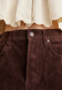 Brown suede pants with a smooth texture, featuring a button closure and subtle stitching details. The waistband sits just below a light blouse.