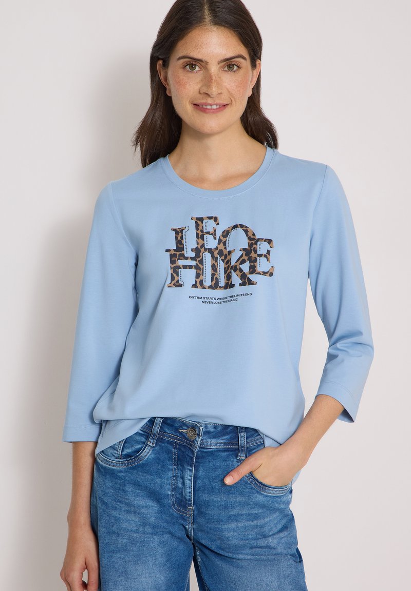 Femme aux longs cheveux foncés portant une chemise bleu clair à manches longues avec un texte imprimé léopard et un jean bleu, debout avec une main dans la poche.