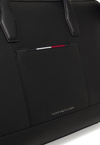 Schwarze Lederhandtasche mit einer strukturierten Oberfläche, ausgestattet mit zwei Griffen, einer Vordertasche mit einem gestreiften Design in Rot, Weiß und Schwarz sowie einem Markenlogo.