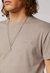 T-shirt color marrone chiaro realizzato in cotone, con maniche corte e logo ricamato "TIGHA" sul petto, arricchito da collane d'argento sovrapposte.