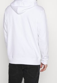 Sweat à capuche blanc en tissu doux, avec une capuche à cordon, des manches longues et des poignets côtelés, associé à un jean foncé.