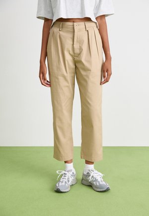 Beige, high-waisted trousers with pleats, cropped length, and a relaxed fit. Paired with light gray sneakers and white socks.
