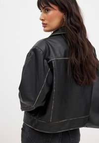 Jauna moteris su ilgais tamsiais plaukais, apsirengusi laisvu juodu odiniu švarku ir juodomis džinsomis, žiūri į kairę prieš paprastą foną.