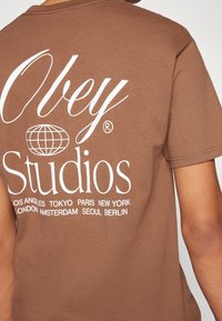 Hnědé bavlněné tričko s bílým textem, logem a grafikou mapy světa na zádech, s krátkými rukávy a kulatým výstřihem.